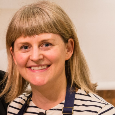 Woman with blond hair smiles at the camera. She is wearing striped shirt and dark apron.