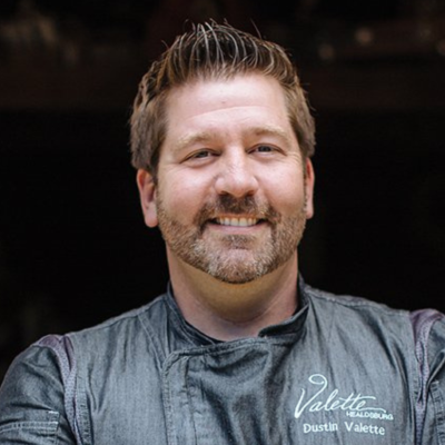 Man with brown hair and beard smiles at camera. He is wearing gray chef's jacket.