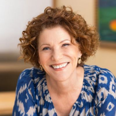 Woman with short curly red hair smiles at the camera. She's wearing a blue and white blouse.