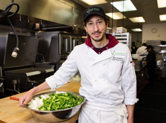 Man in chef coat and black baseball cap stands in kitchen with bowl of greens.