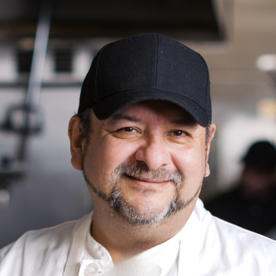 community-class-chef-luis-realpozo-sq Man with short beard and black baseball cap wears white chef's coat and apron