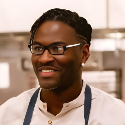 Black man with short hair and glasses. dressed om wjote sjort amd blue apron