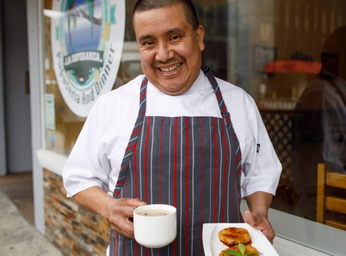 Man in white shirt and striped apron holds plate of food in front of restaurant door