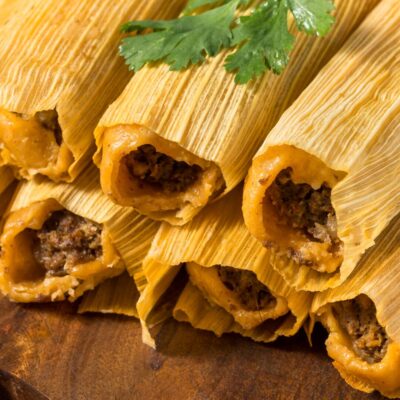 Tamales wrapped in corn husks stacked on a board with cilantro